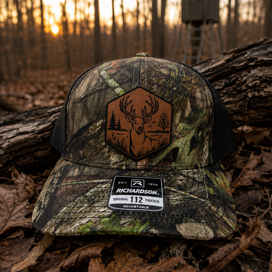 A camouflage trucker hat with a brown leather patch featuring a deer graphic, resting on a log in a forest during sunset. The hat has a black mesh back and a Richardson 112 Mossy Oak Country DNA front.